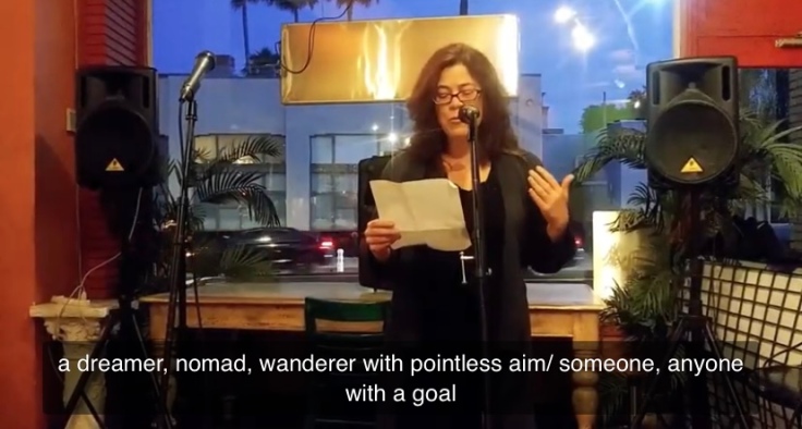 screenshot of me standing behind a microphone in front of a window at a coffee shop. I am holding a piece of paper reading a poem. Text on screen says a dreamer, nomad, wanderer with pointless aim/ someone, anyone with a goal.